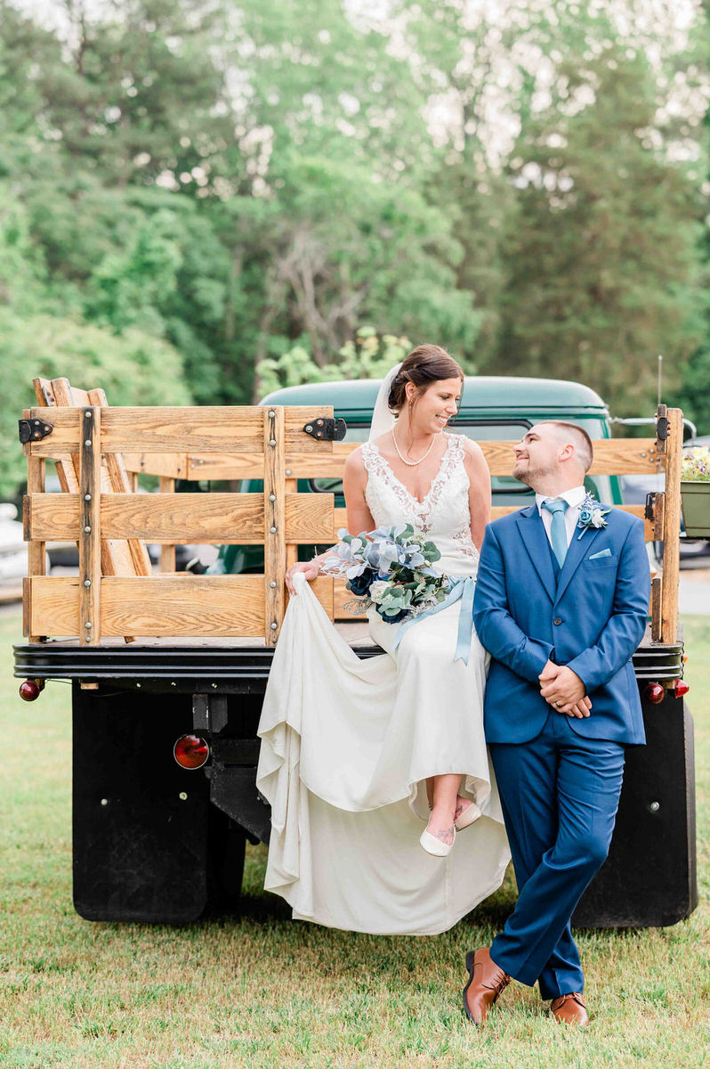 A bride sitting on an old truck while a groom stands next to her and looks at her at one of the best outdoor wedding venues in Richmond VA.