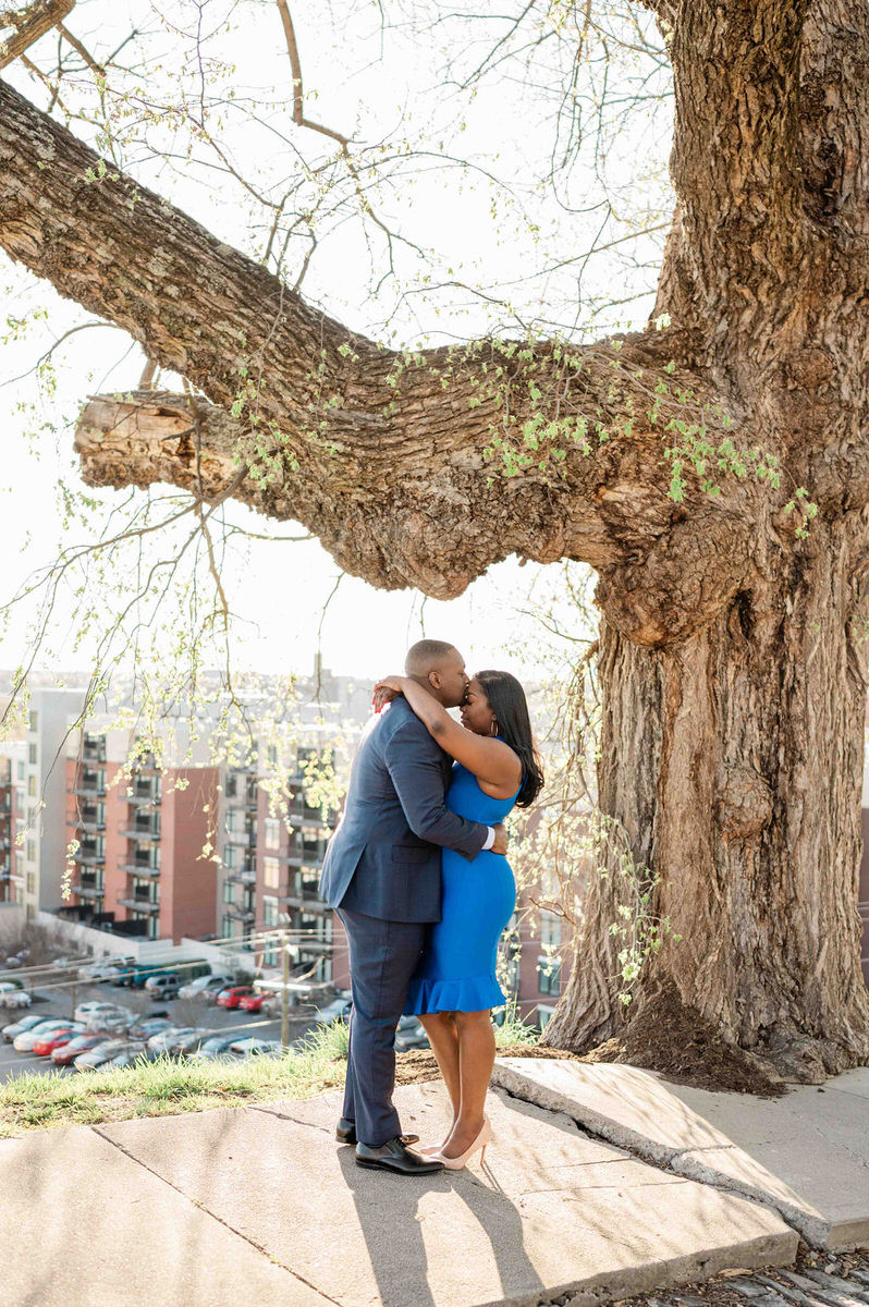 A couple in semi-formal attire sharing a kiss under a large tree, with a cityscape spread out behind them in the soft glow of sunlight in one of the best places to take pictures in Richmond VA.