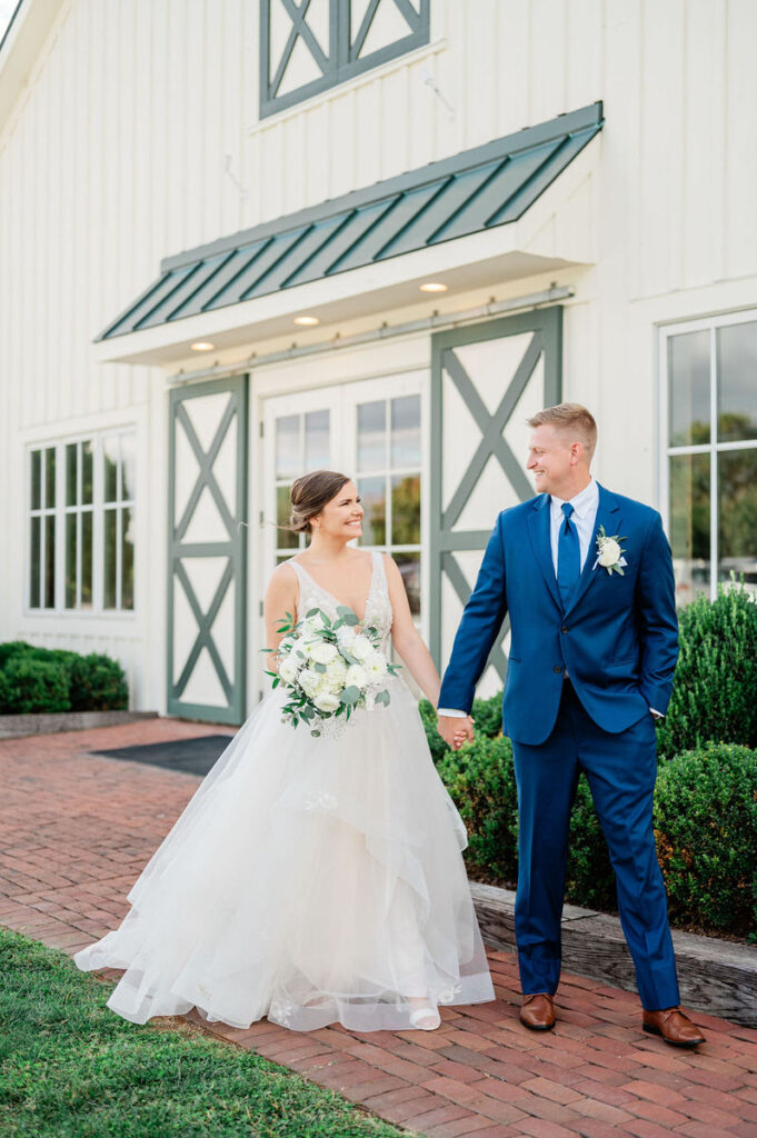 A bride and groom smiling at each other in front of a white barn with green shutters, with the bride holding a bouquet of white flowers