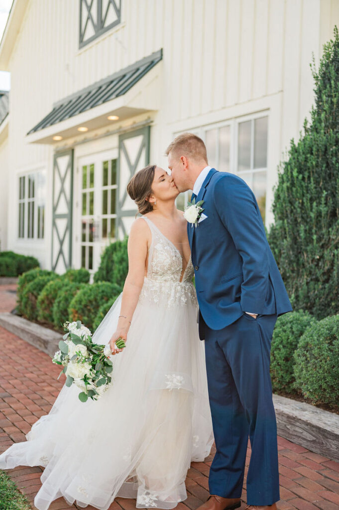 A newlywed couple kissing, with the bride holding a bouquet and the groom in a blue suit, standing in front of a white barn with green shutters