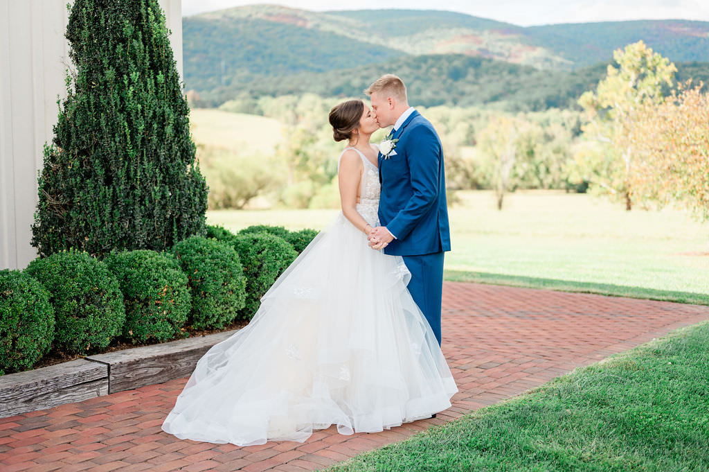 A bride and groom kissing on a brick pathway, framed by lush greenery and mountain views in the background.