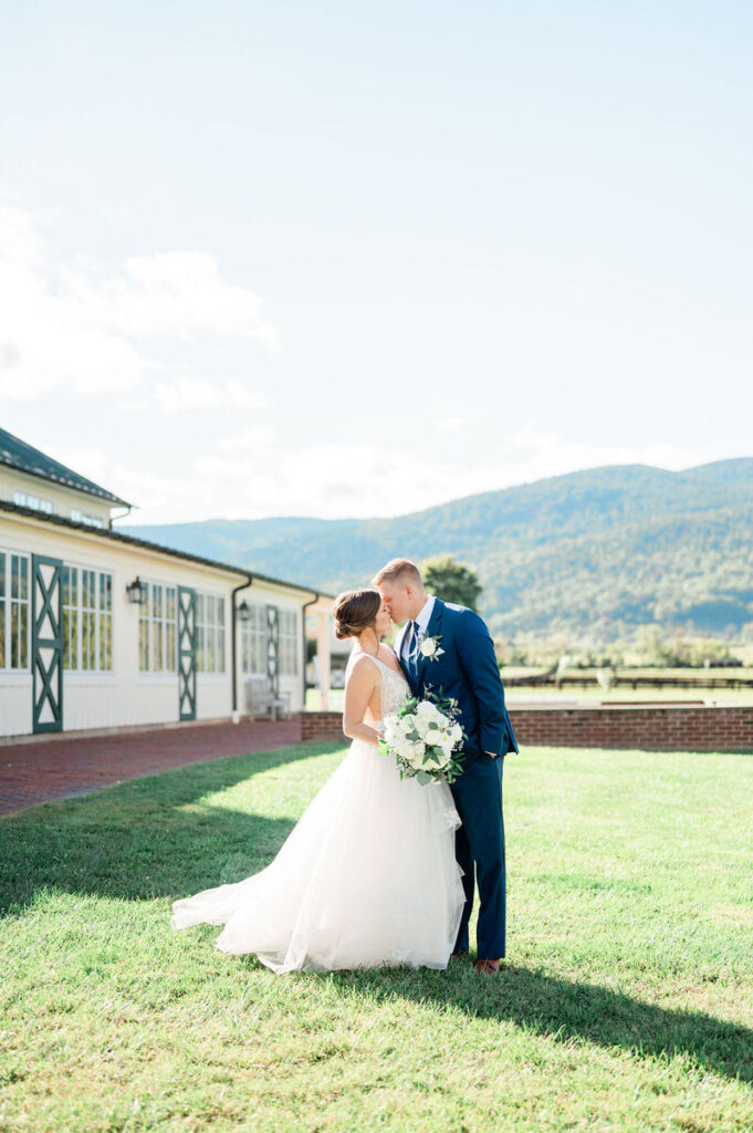 A bride and groom sharing a kiss on a lawn with a majestic mountain backdrop, near a white building with green trim