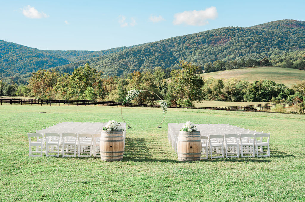 An outdoor wedding ceremony setup at the King Family Vineyards wedding venue featuring white chairs arranged around a circular floral arch, set in a field with mountains in the distance