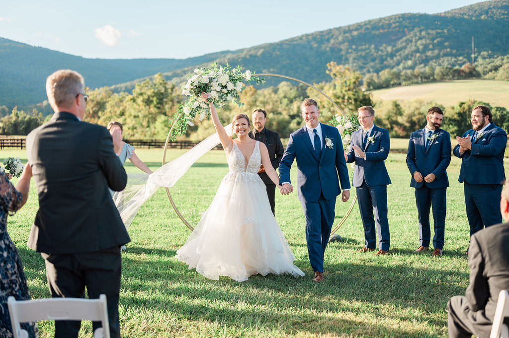 A bride triumphantly raises her bouquet as she and the groom walk down the aisle, while guests applaud, with mountains under a clear blue sky behind them.

