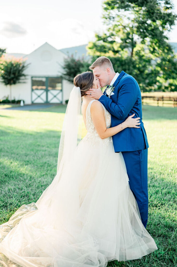 A bride and groom embrace and share a kiss on a lawn, with the bride's veil flowing and the groom's blue suit complementing the clear sky.

