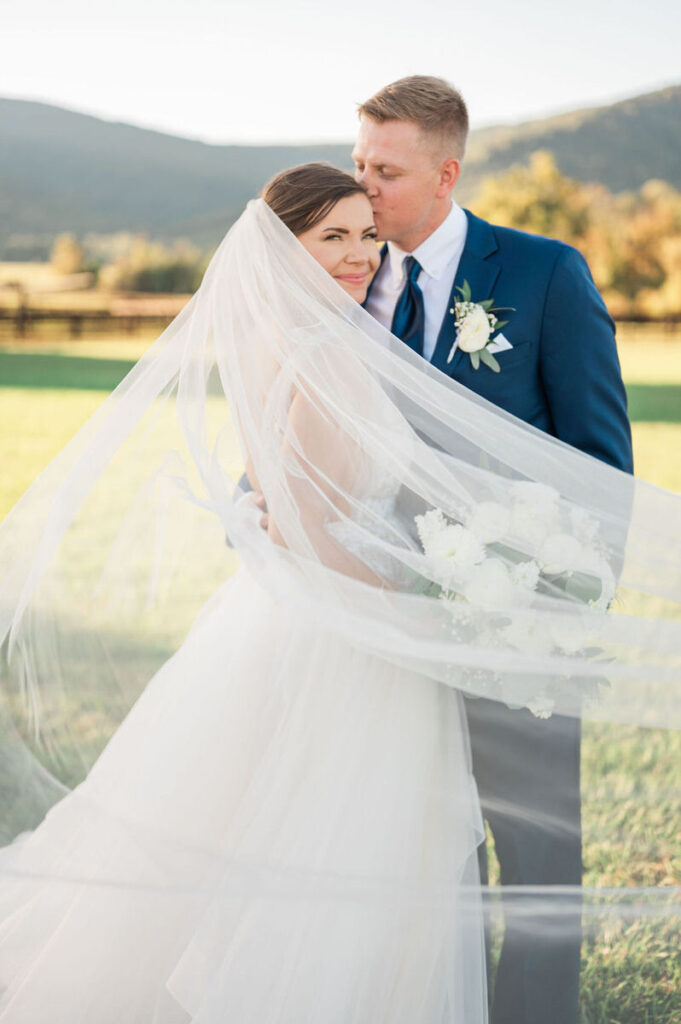 A bride and groom wrapped in the bride's veil, sharing an intimate moment with mountain scenery in the background.

