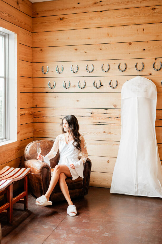 A bride sitting in a leather armchair, holding a champagne flute, with a white wedding dress hanging beside her against a wooden wall adorned with horseshoes