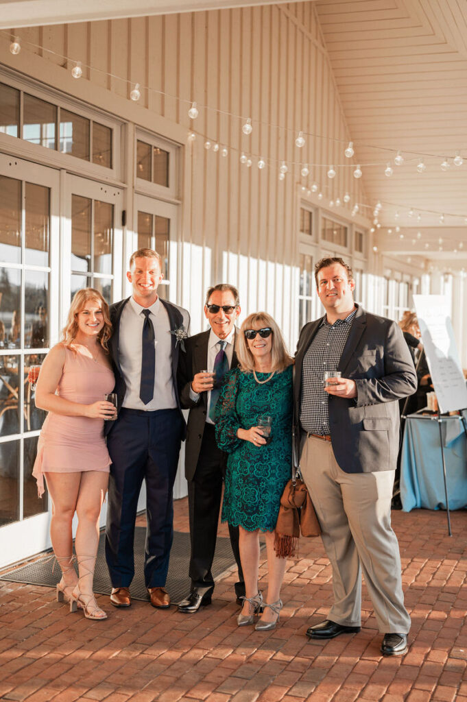 Guests at a wedding reception standing on a porch, with a man in a blue suit taking a photo with three others, under a string of lights.

