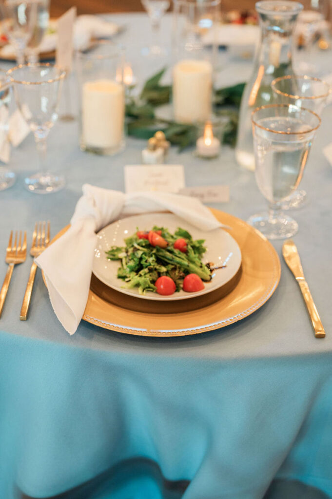 A wedding dinner place setting with a blue tablecloth, gold-rimmed plates, and a fresh salad, presenting an elegant meal arrangement.

