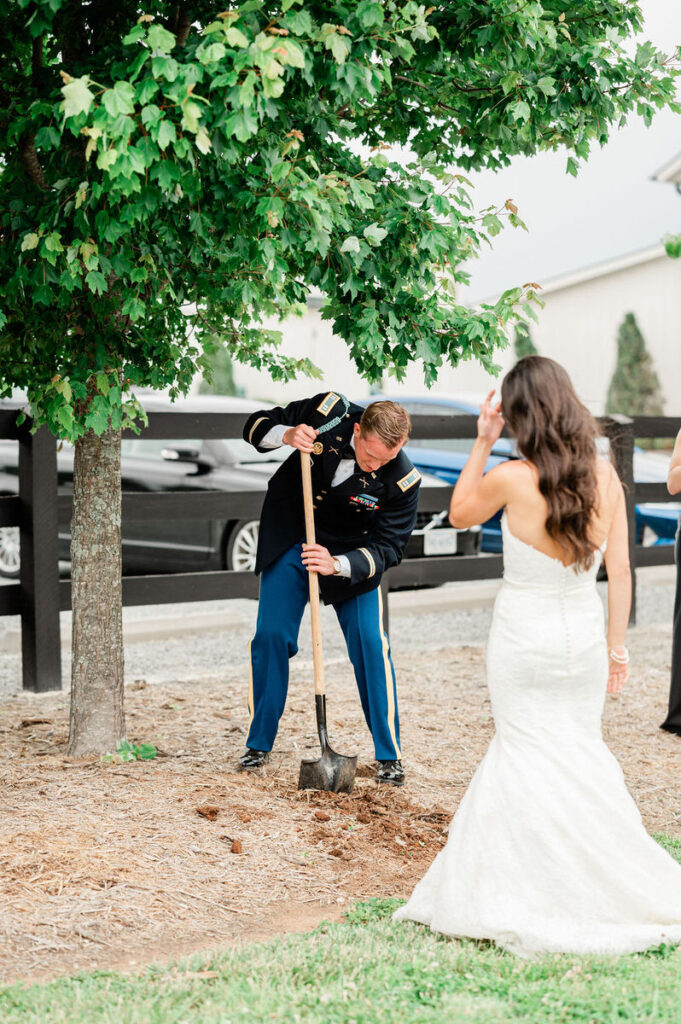 A man in a military uniform digging with a shovel next to a bride, performing a tree-planting ceremony