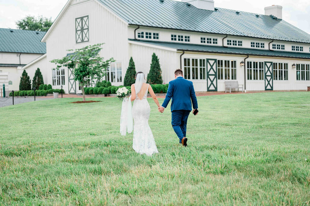 A couple holding hands and walking away across a lush lawn, with the back of the bride's lace dress and the groom's blue suit in focus