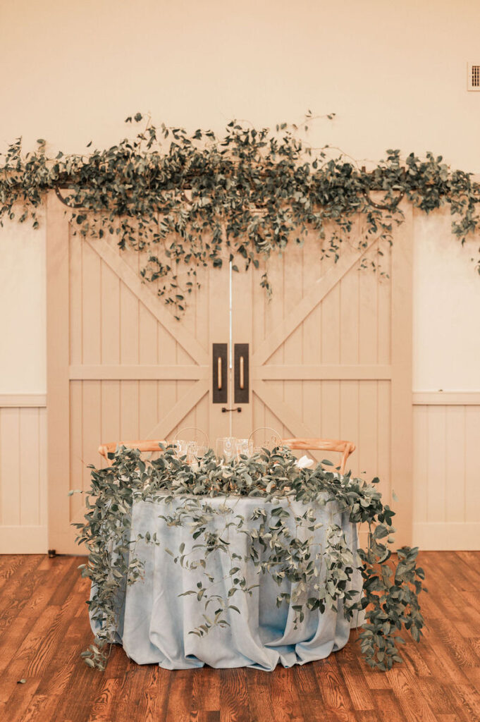 An elegantly decorated wedding sweetheart table with a cascading eucalyptus garland over a pale blue tablecloth, set against wooden barn doors