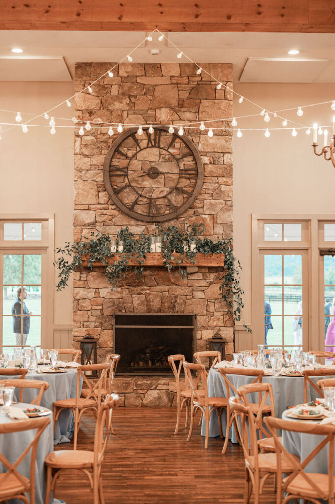 A rustic-chic wedding reception setting featuring a stone fireplace with a decorative greenery mantle, under string lights, and surrounded by wooden chairs and tables with blue tablecloths