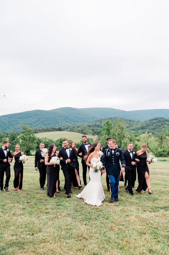 A bridal party walking and laughing together across a grassy field, with the bride and groom in the center, surrounded by groomsmen in black suits and bridesmaids in black dresses