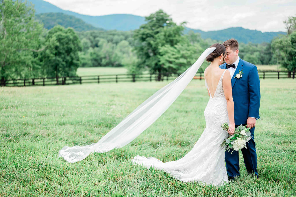 A bride and groom sharing a romantic moment in a field, with the bride's veil trailing in the wind and the groom in a blue suit holding a bouquet