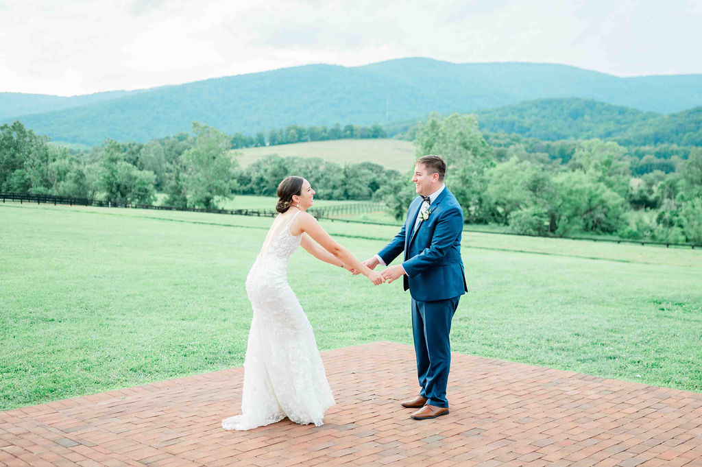 A couple holding hands and dancing on a brick patio, set against a backdrop of rolling hills and a verdant landscape