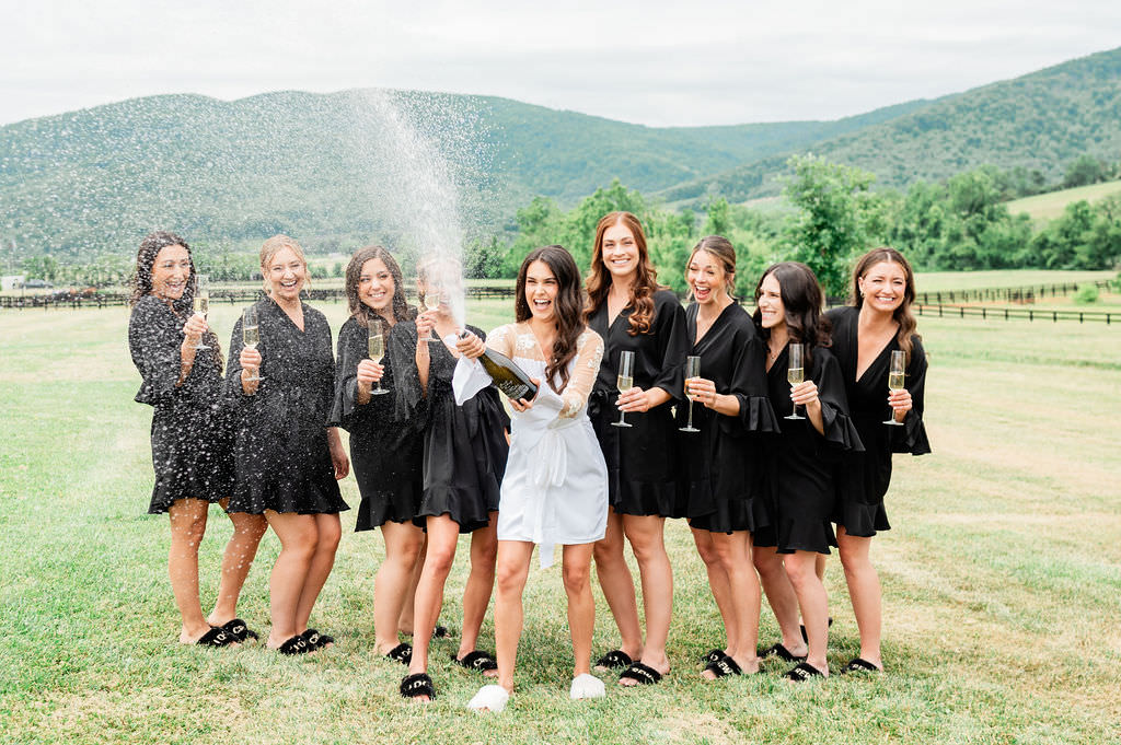 A group of joyful bridesmaids popping champagne and celebrating, with one in a white dress opening the bottle, and the others in black dresses laughing and holding glasses, all set against a backdrop of rolling hills and a fenced pasture.