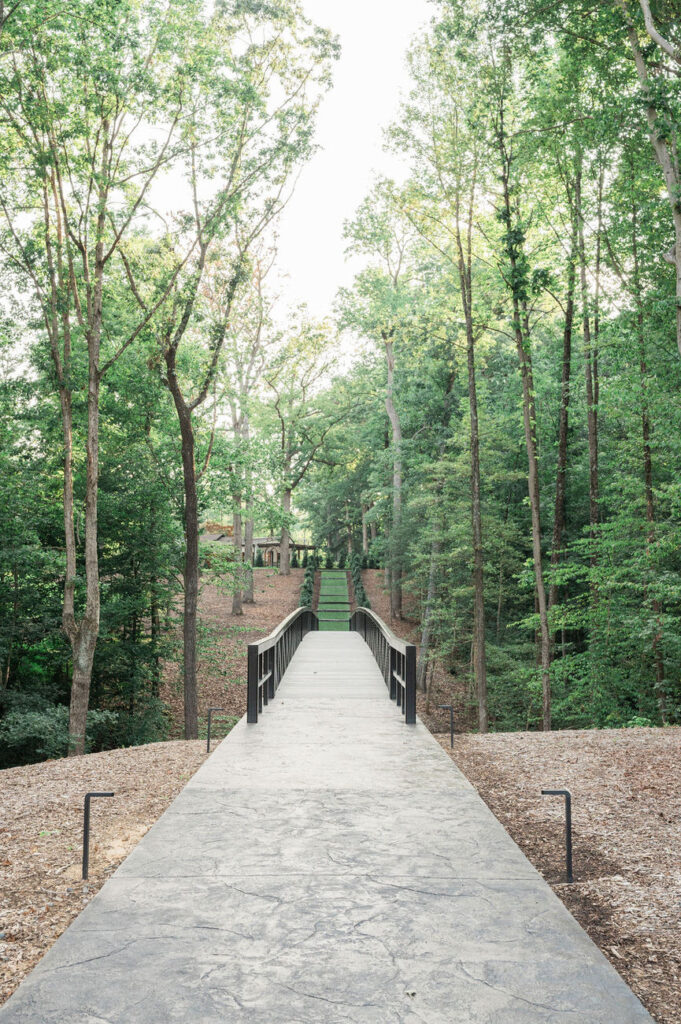 A serene pathway leading over a bridge surrounded by lush greenery, creating a tranquil nature walk within the Hidden Oaks venue.