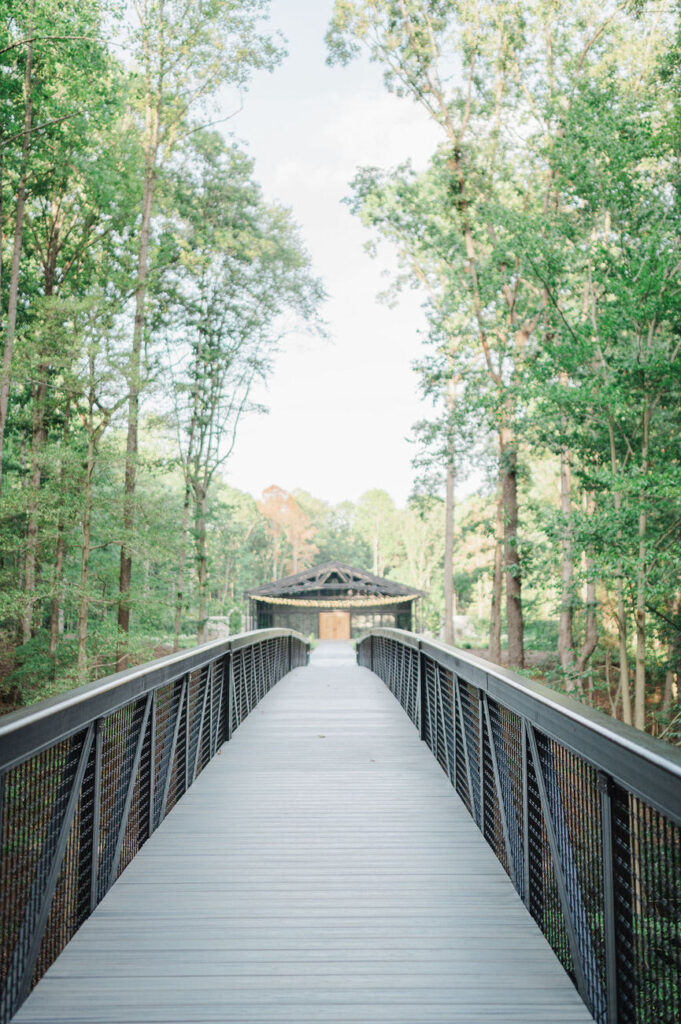 A long, elevated walkway framed by manicured shrubbery and tall trees leading to a covered pavilion at the Hidden Oaks venue, set in a tranquil forest setting.