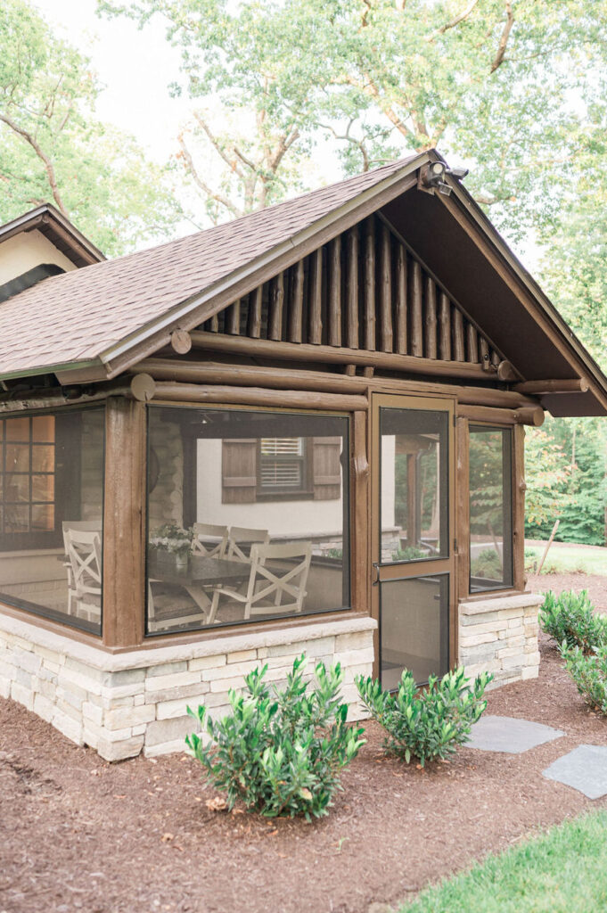 A close-up view of a quaint cottage at Hidden Oaks venue, highlighting the log cabin design, screened porch area with seating, and a landscaped garden with young shrubs.