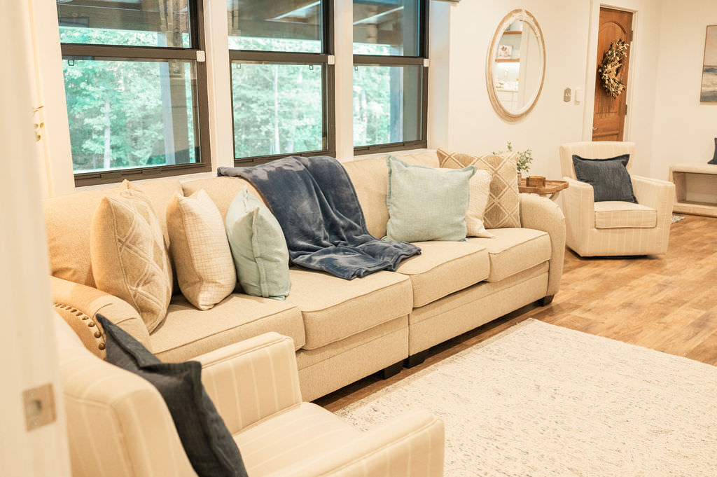 A cozy living room with a cream sofa filled with decorative pillows, a matching armchair, and large windows allowing for ample natural light, set in a warm, inviting color scheme.