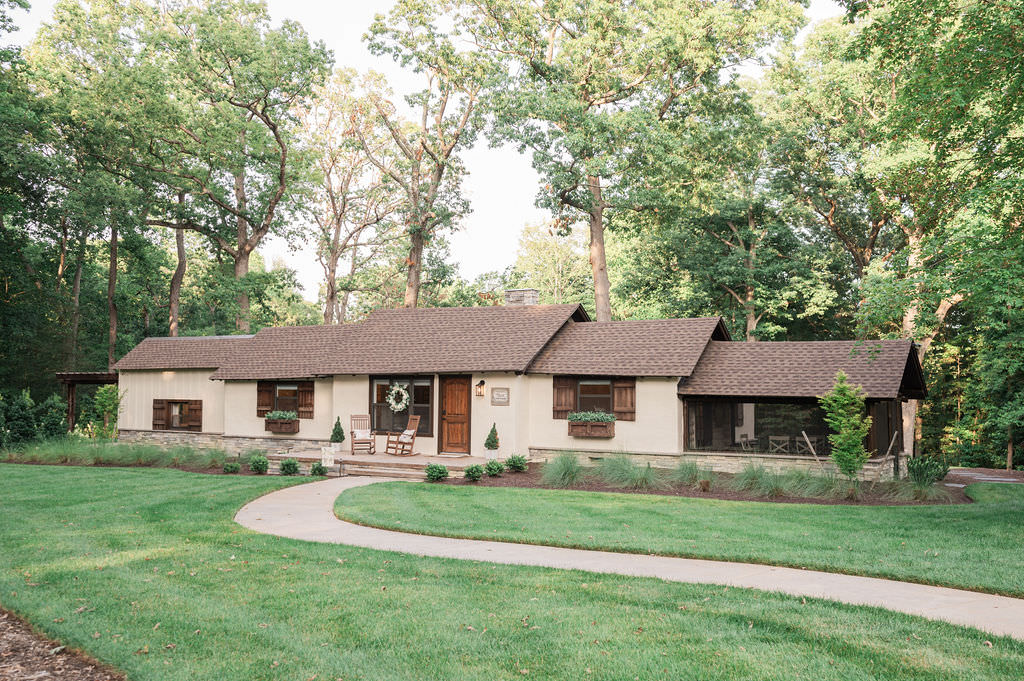 A charming single-story cottage at Hidden Oaks venue, with beige walls and a brown roof, nestled among mature trees and manicured lawns, featuring a welcoming front porch.