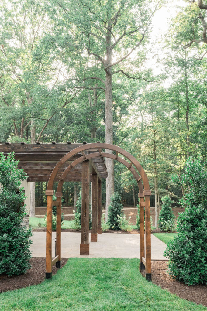 A wooden archway at the Hidden Oaks venue providing an entrance to a paved area surrounded by lush greenery, creating a picturesque outdoor ceremony or event space.