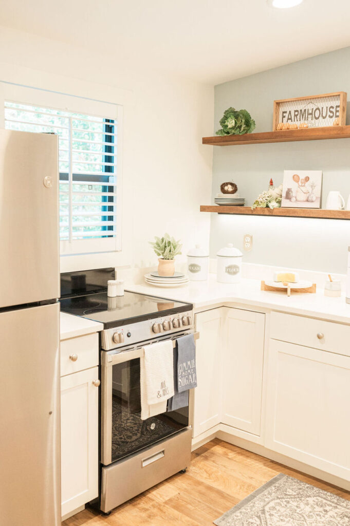 A bright, farmhouse-style kitchen with white cabinets, open shelving displaying decorative items, and modern appliances, giving off a clean and homey vibe.