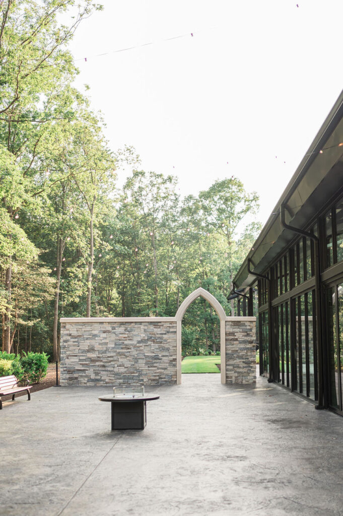 A serene outdoor patio featuring a stone firepit table, benches, and lush greenery, complemented by the modern architecture of the venue with large windows and an arched doorway.