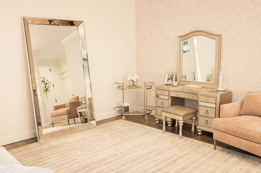 A spacious dressing room featuring a large full-length mirror on the left, a classic wooden vanity set with a matching stool, and a comfortable couch in the corner, all set against a patterned wallpaper.