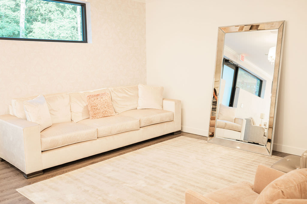 A minimalist and chic getting ready space with a large off-white sofa adorned with decorative cushions, a matching armchair, and a full-length mirror reflecting the light-filled room.