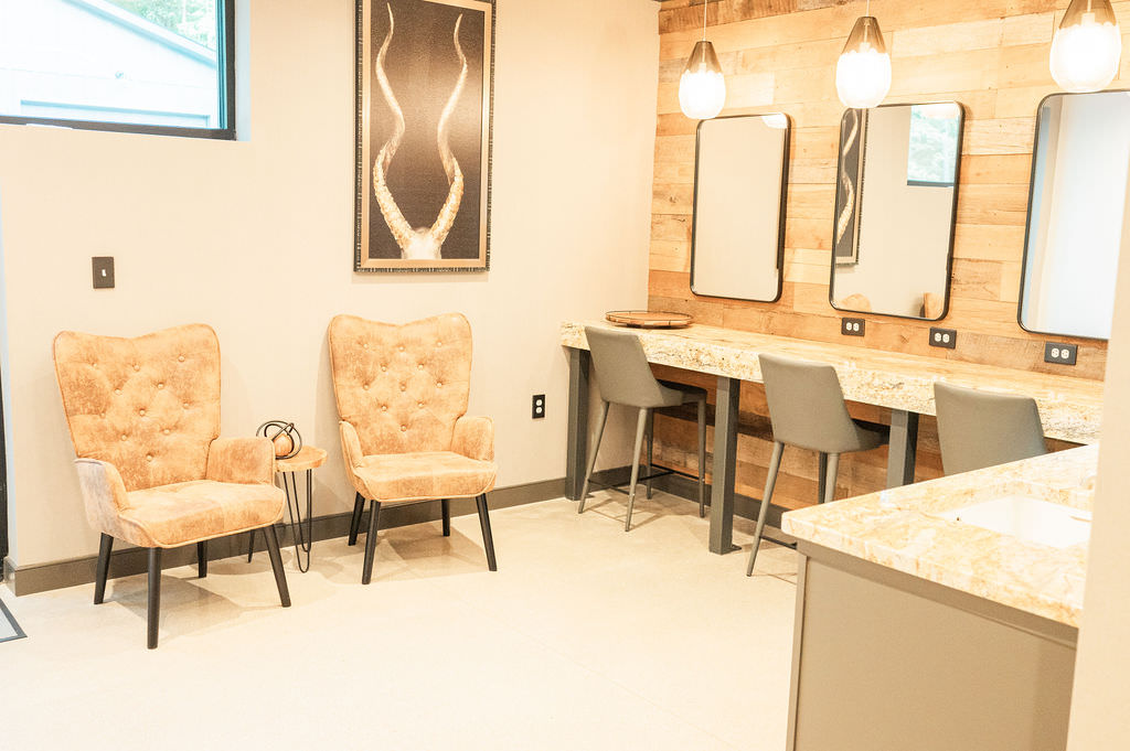 A bright and airy bridal suite setup with two luxurious velvet chairs, a long counter with built-in mirrors and modern pendant lights, creating a welcoming space for bridal preparations.