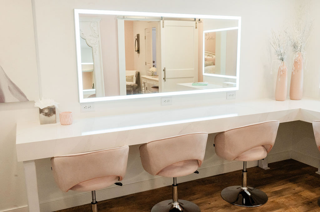 A modern and elegant vanity area with a large mirror surrounded by bright lights, two plush pink chairs, and a marble countertop. The reflection shows a glimpse of the adjoining room.