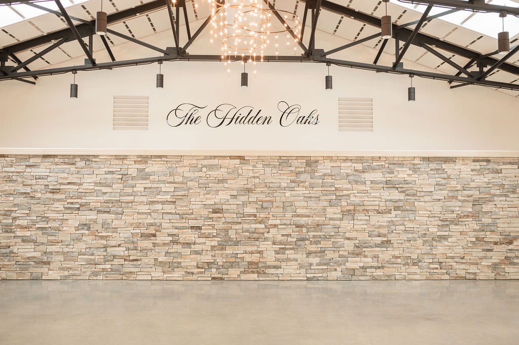 A wide shot of the reception hall showcasing 'The Hidden Oaks' wall signage above a stone feature wall, with elegant circular chandeliers providing soft lighting to the spacious area.