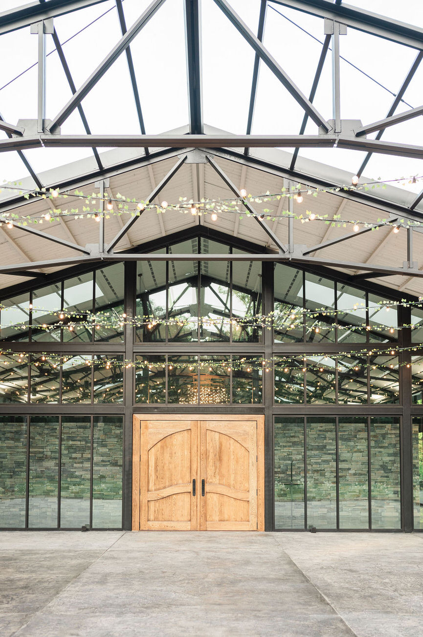 The entrance to a greenhouse reception area at Hidden Oaks venue, with a striking wooden double door, large glass panels, and fairy lights overhead, creating a modern yet rustic entryway.
