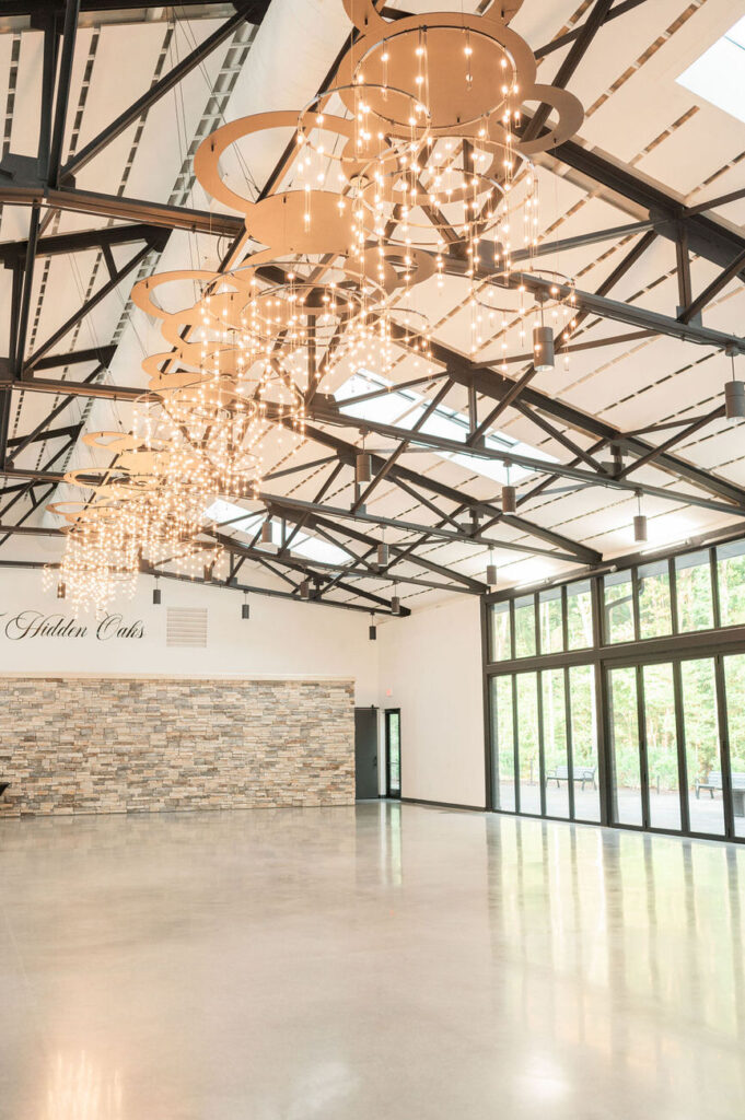 Another perspective of the reception hall highlighting the grandeur of the space with its high ceiling, adorned with unique circular chandeliers and fairy lights, complemented by the stone wall and large windows.






