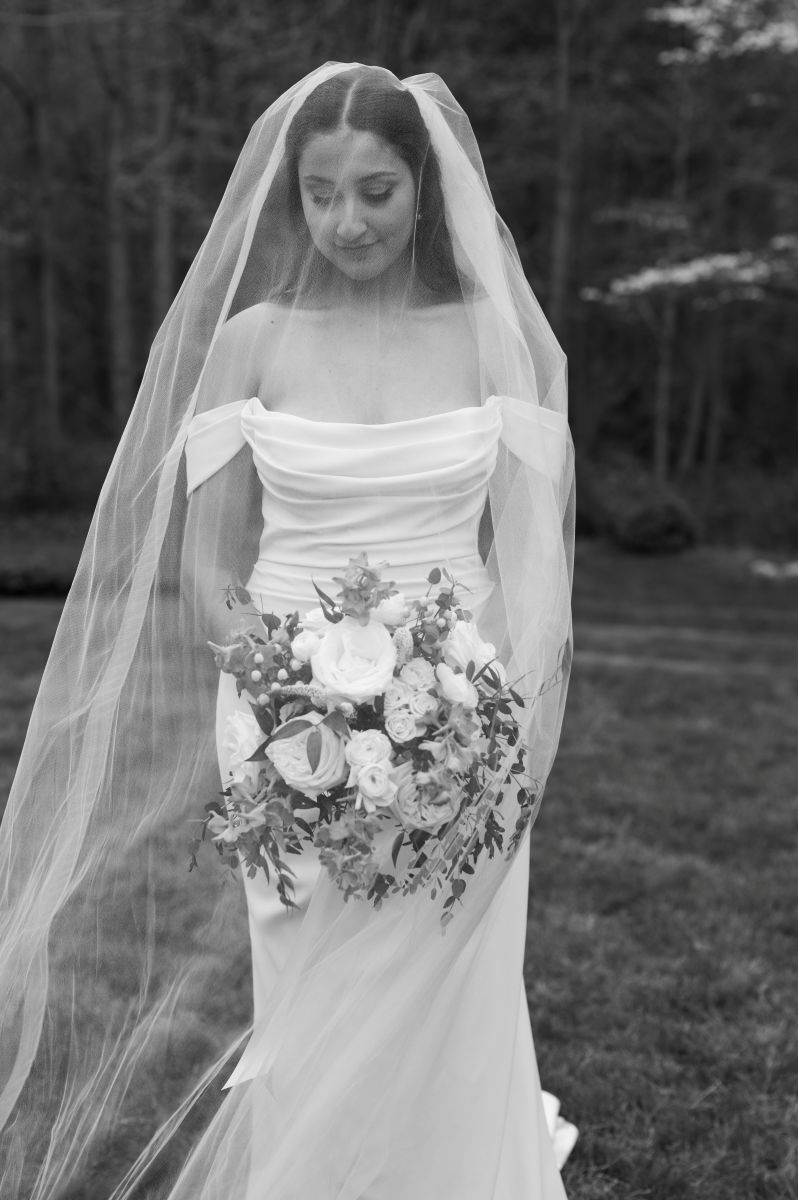 a woman wearing a white wedding dress and veil standing outside holding a bouquet of flowers