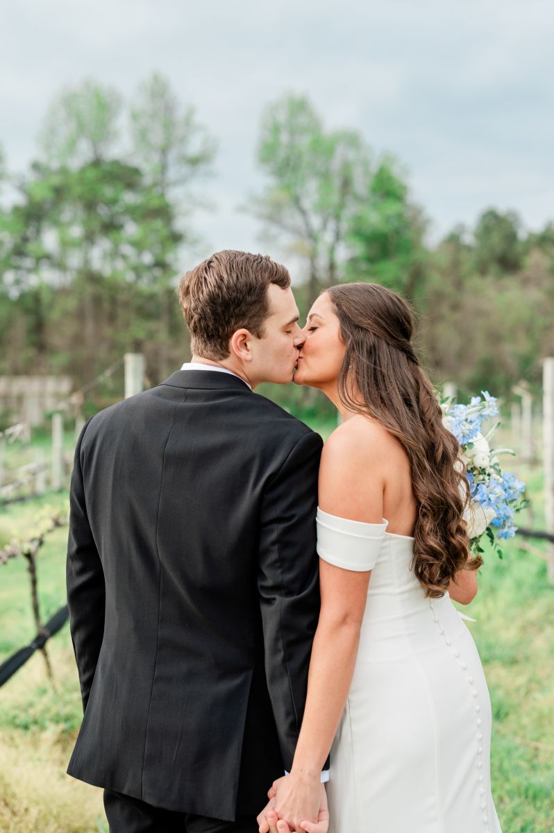 a couple kissing in the vineyards at ashton creek vineyard in virgina the woman is wearing a white wedding dress and holding flowers the man is wearing a black suit