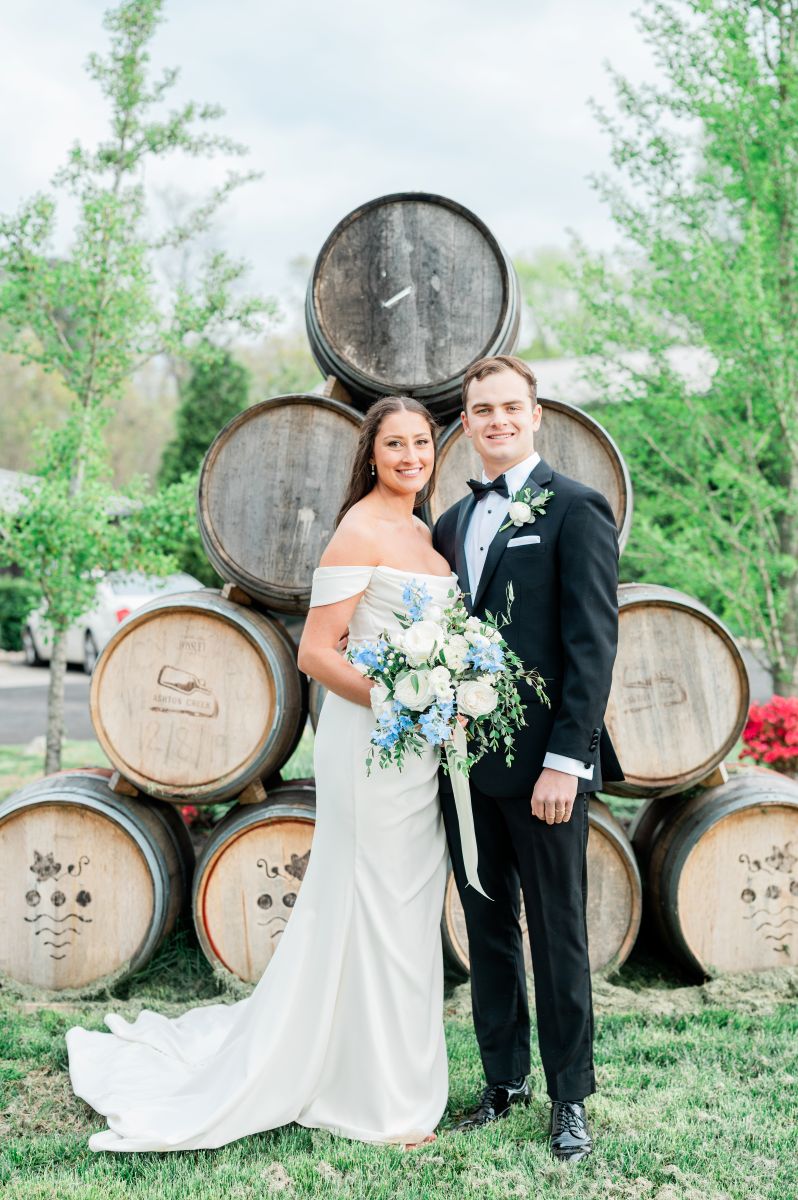 a couple smiling and standing in front of wine barrels at a vineyard the woman is wearing a white wedding dress and holding flowers the man is wearing a black suit