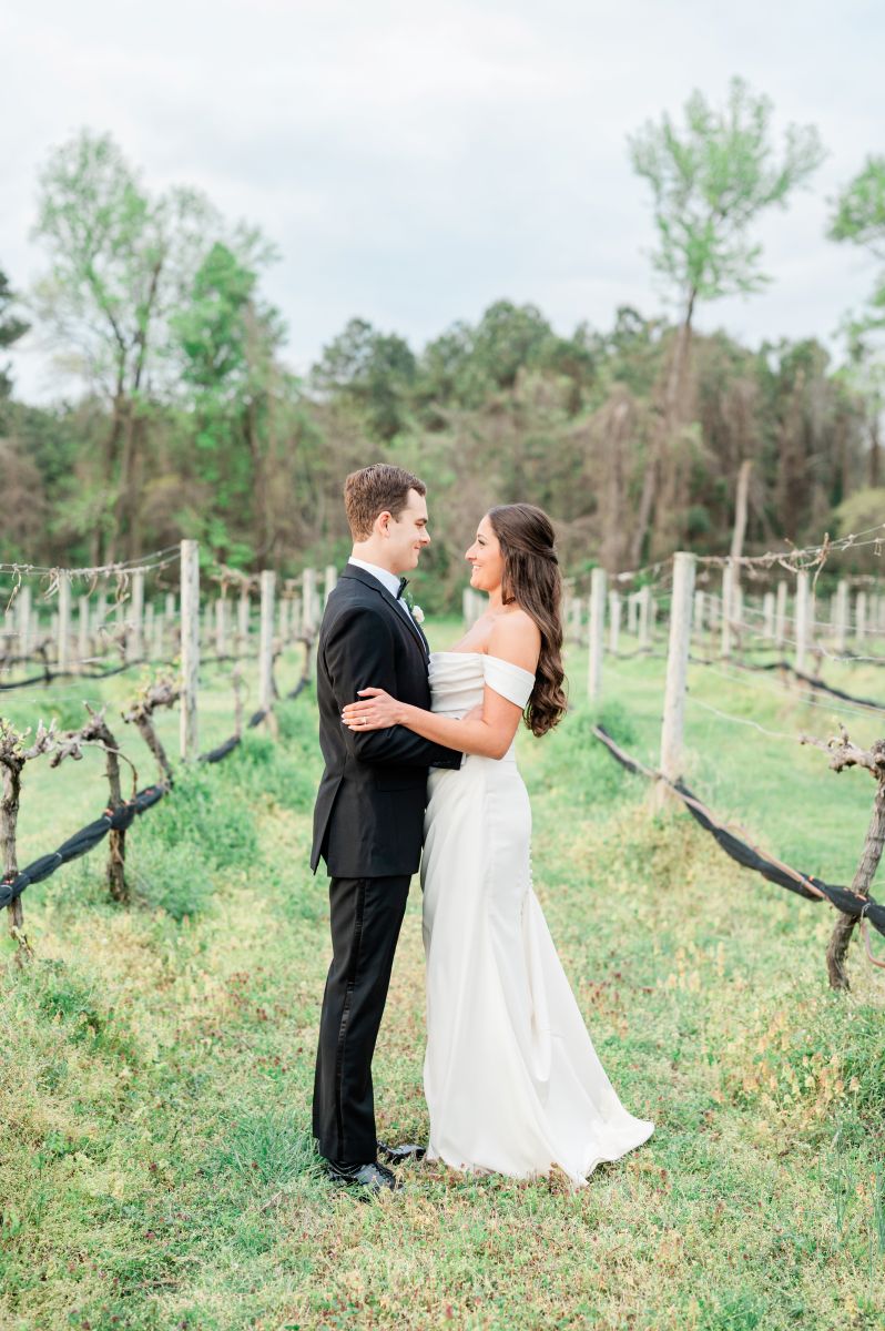 a couple embracing in the vineyards at ashton creek vineyard they are smiling at each other and the woman is wearing a white wedding dress the man is wearing a black suit