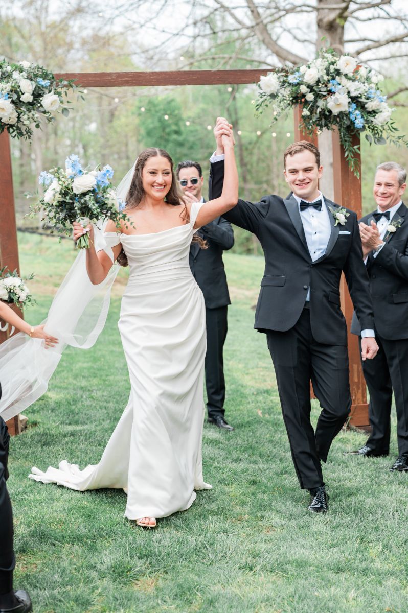 a couple leaving their wedding ceremony at ashton creek vineyard they have their hands in the air and are smiling at each other the man is wearing a suit the woman is wearing a white wedding dress and veil and holding a bouquet of flowers
