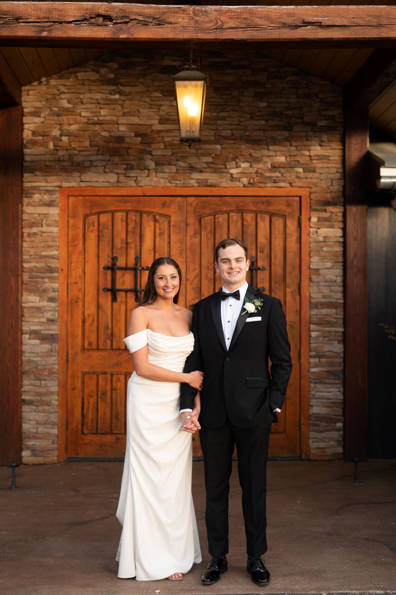 a couple standing in front of a barn door smiling the woman is wearing a white wedding dress the man is wearing a black suit