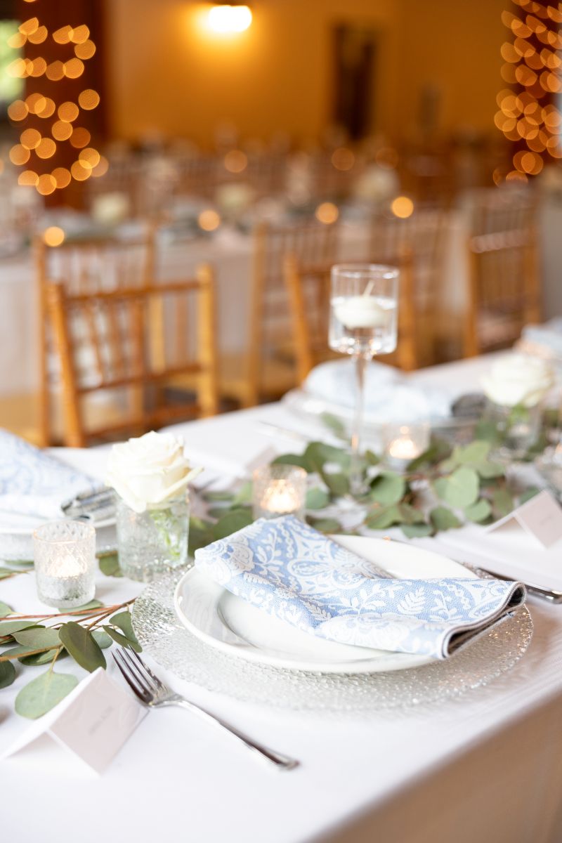 a banquet table adorned with a white tablecloth, plates, eucalyptus, glasses, and candles set up for a wedding reception