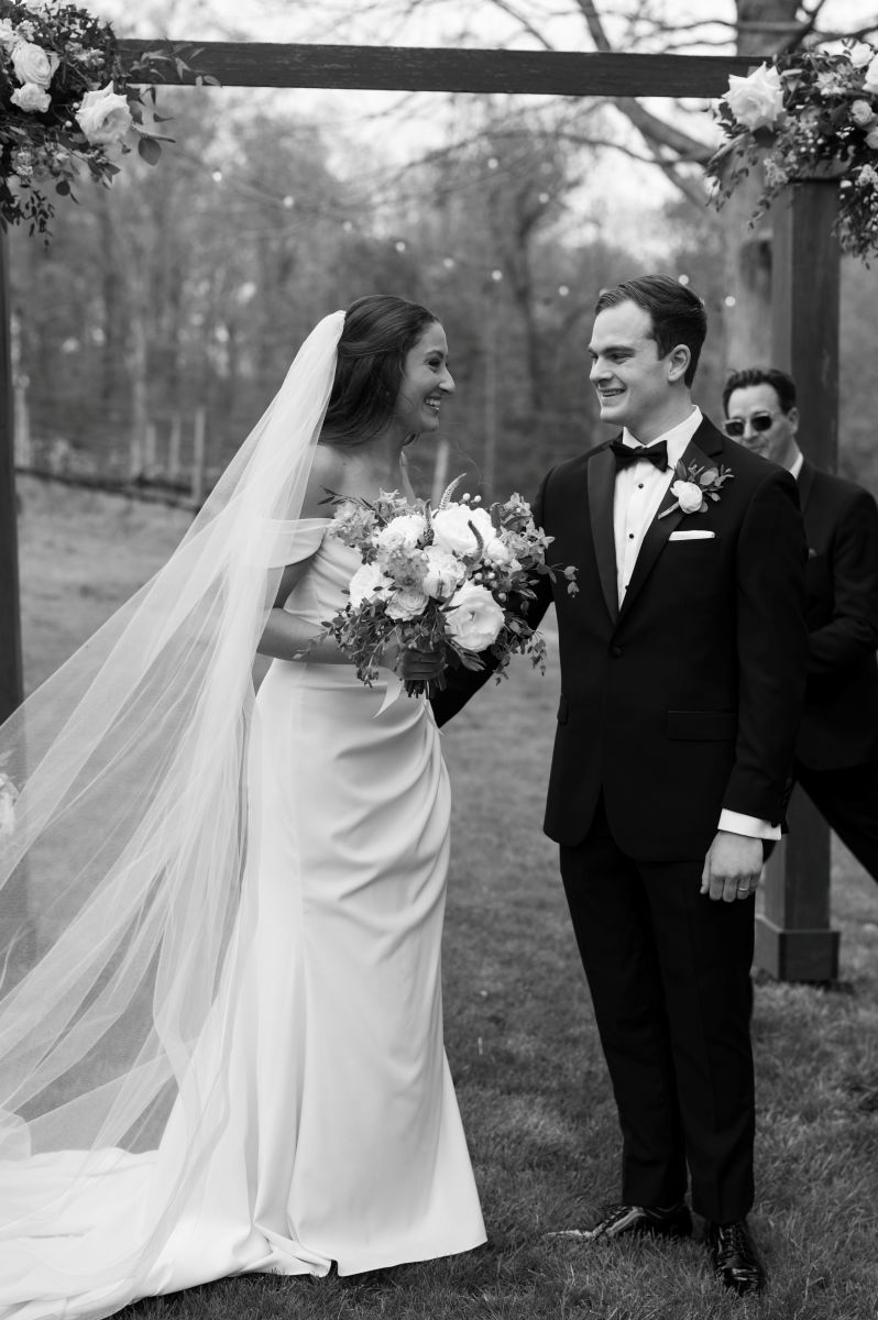 a copule laughing together after their wedding ceremony the woman is wearing a white dress and veil and holding flowers the man is wearing a black suit