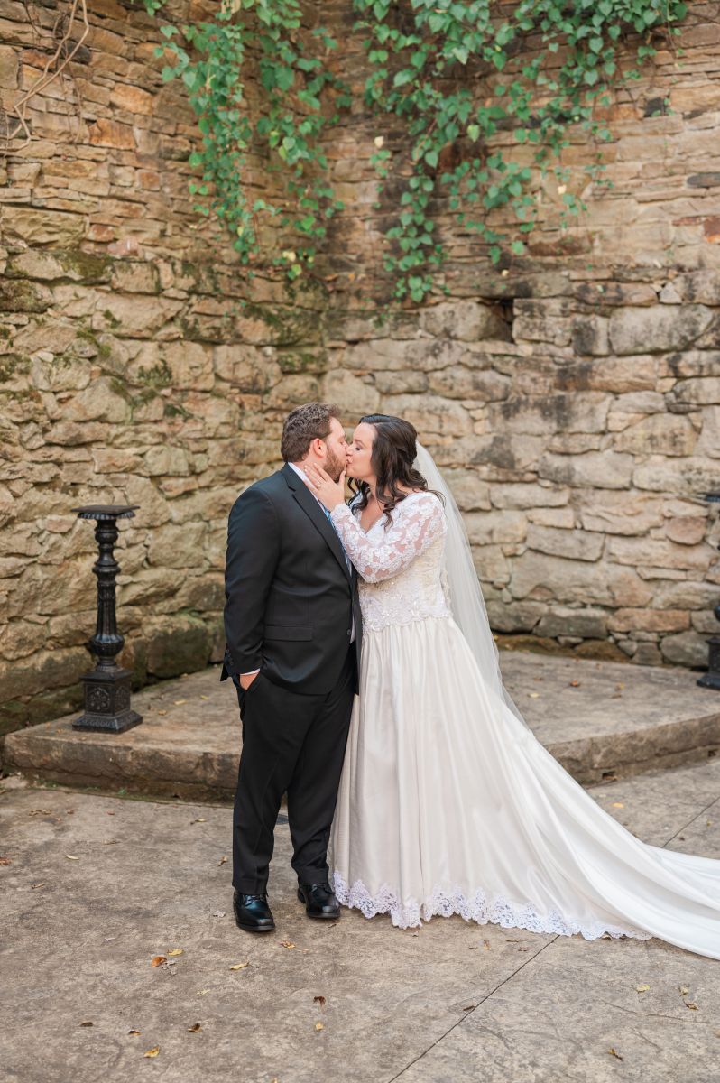 a woman wearing a white wedding dress and veil is kissing her partner at the ceremony site called the ruins at The Mill at Fine Creek in richmond virginia