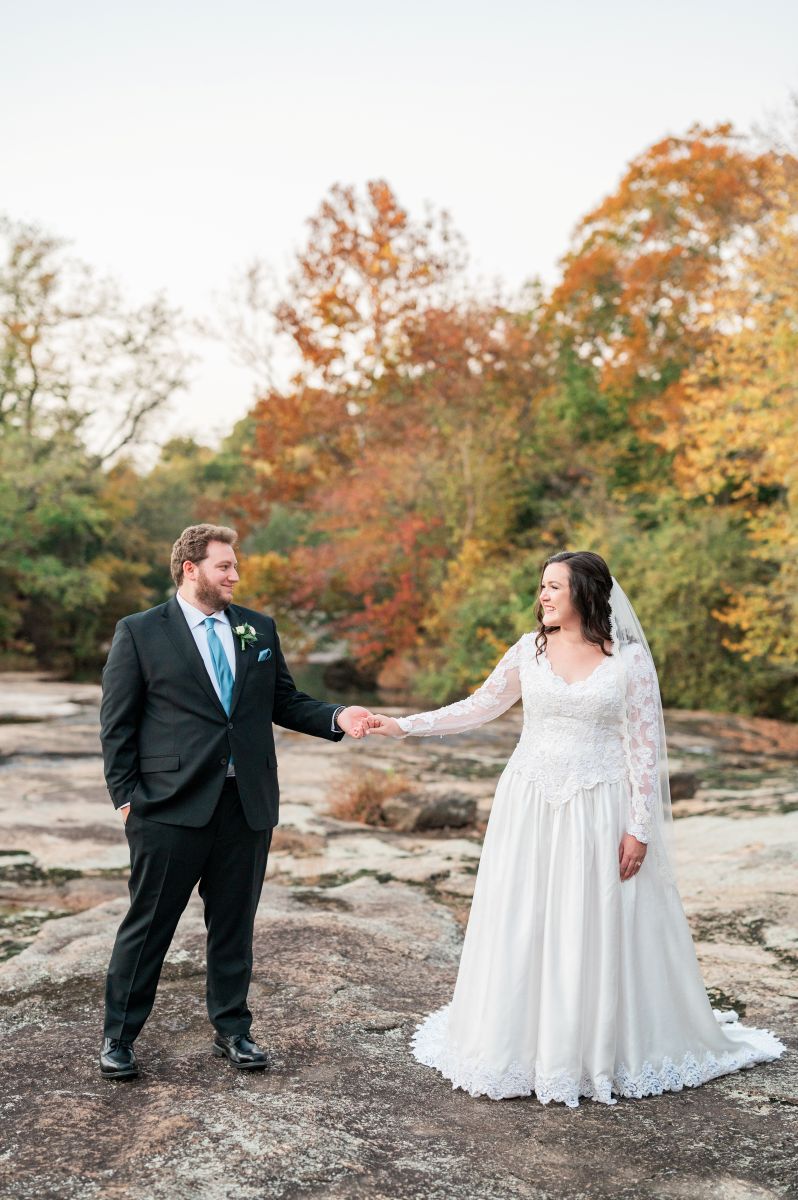 a copule standing on rocks next to trees holding hands and smiling at each other the woman is wearing a white wedding dress and veil the man is wearing a black suit
