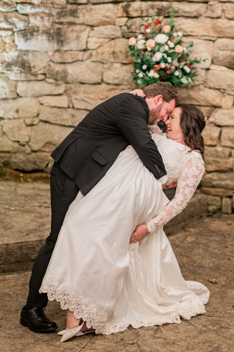 a man wearing a black suit dipping his partner backwards who is wearing a white wedding dress and heels they are standing in the ruins ceremony site at the mill at fine creek wedding venue in richmond virginia