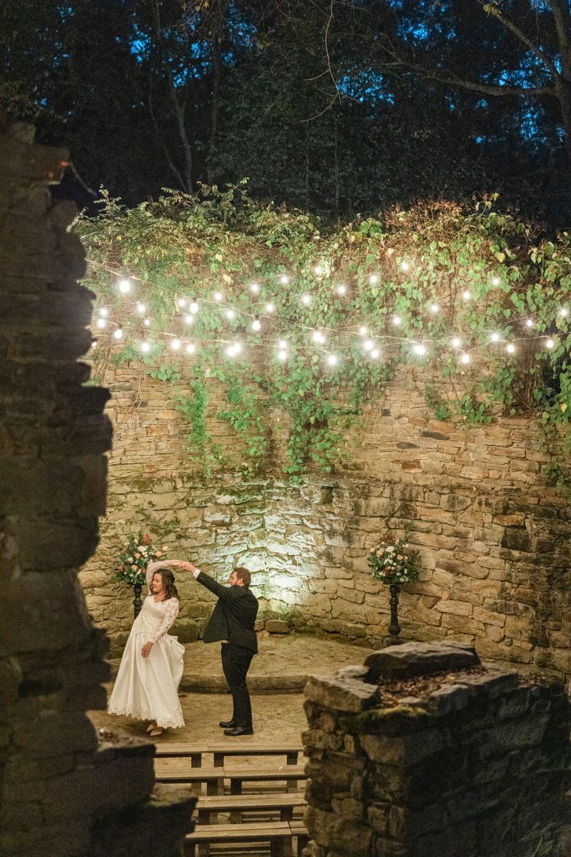 a couple dancing in the ruin ceremony site underneath twinkling lights at the mill at fine creek in richmond virginia the woman is wearing a white wedding dress the man is wearing a black suit
