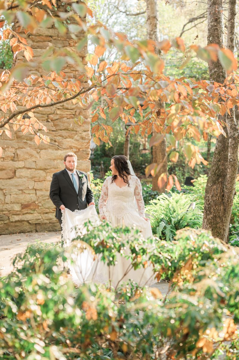 a couple walking down a path during the fall the woman is wearing a white wedding dress and veil and turns to look back at her partner who is wearing a black suit and helping carry her wedding dress