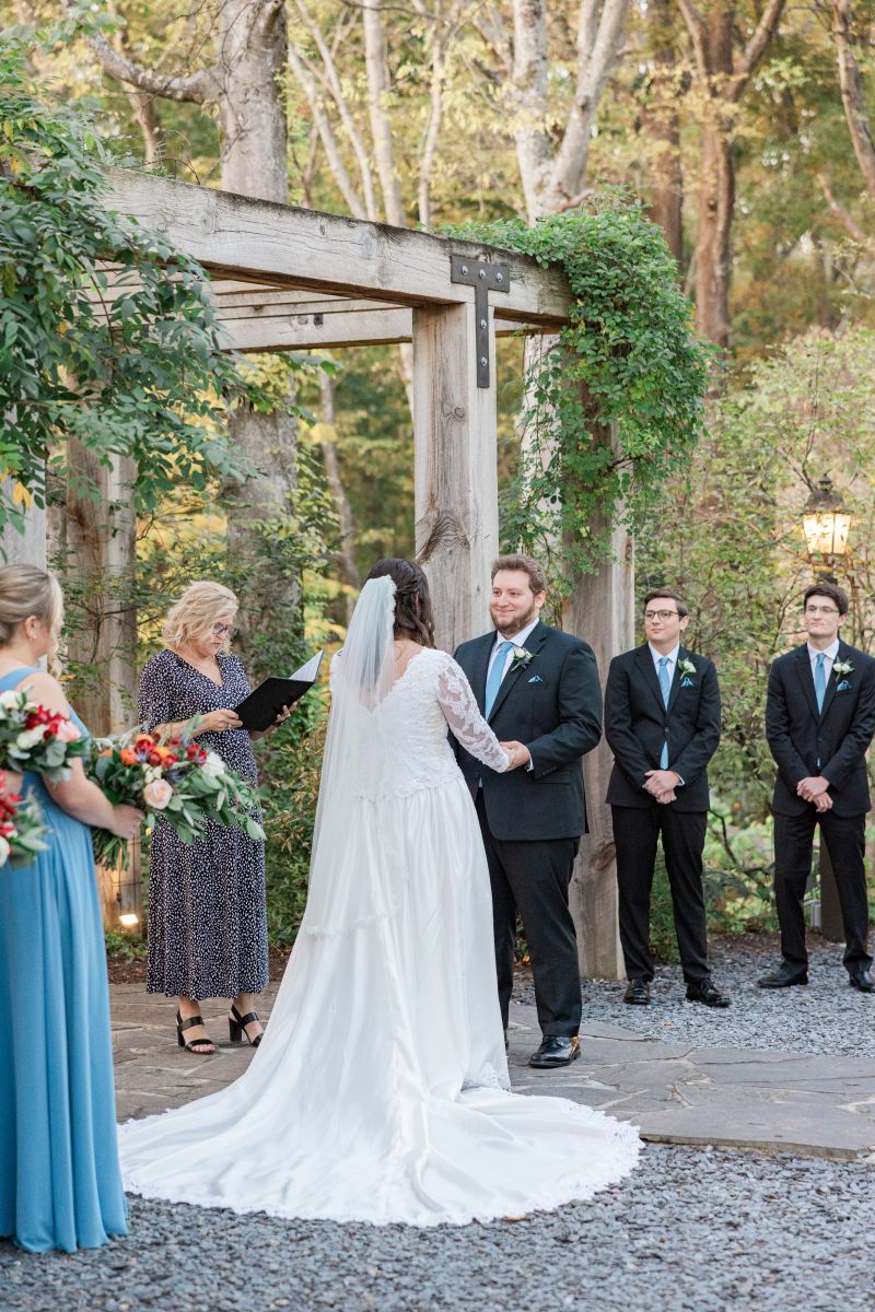 a couple at their wedding ceremony holding hands and looking at each other the woman is wearing a white wedding dress and veil the man is wearing a black suit their wedding party is next to them as well as their officiant